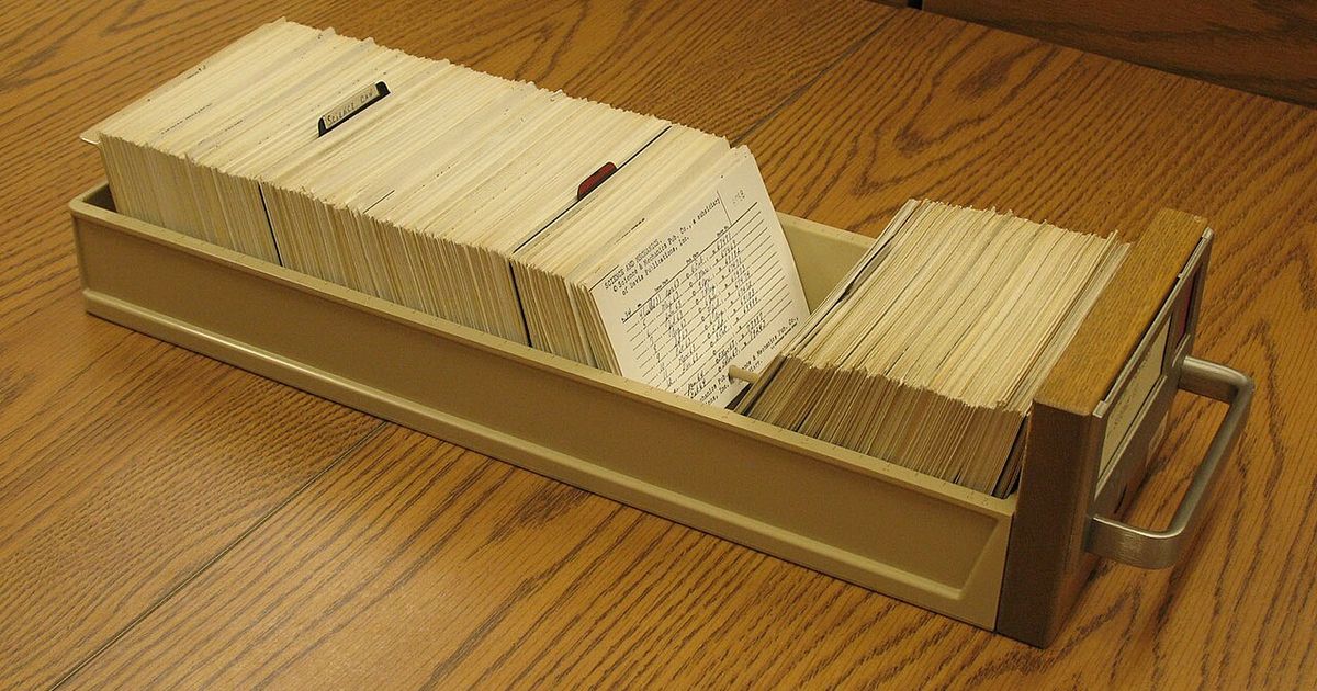 A beige tray of index cards on a wooden table, with one card at the front showing handwritten entries