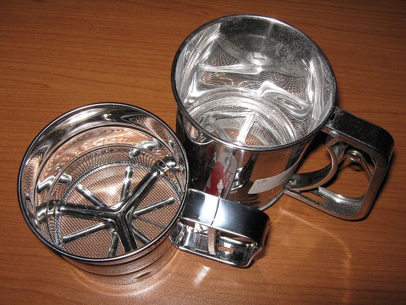 Two stainless steel flour sifters on a wooden table, one turned over to show its mesh